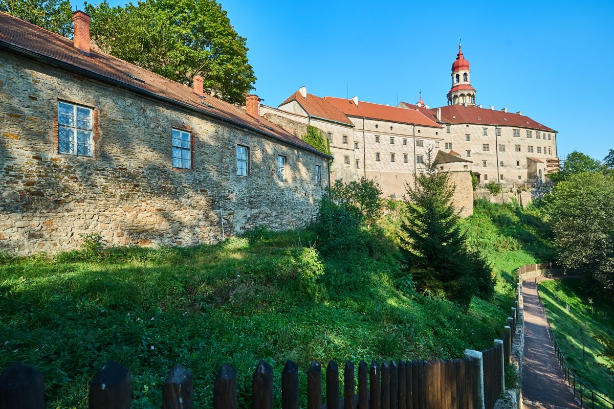 Panorama wzgórza z Zamkiem w Náchodzie, otoczonym murami obronnymi i zielenią. Widać kamienne zabudowania z czerwonym dachem i wieżę z charakterystyczną czerwoną kopułą.