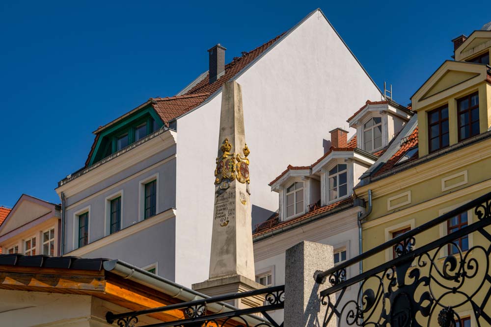 Słup dystansowy poczty polsko-saskiej w Zgorzelcu stojący pomiędzy kamienicami o pastelowych elewacjach, widoczny w pełnej wysokości w popołudniowym słońcu.