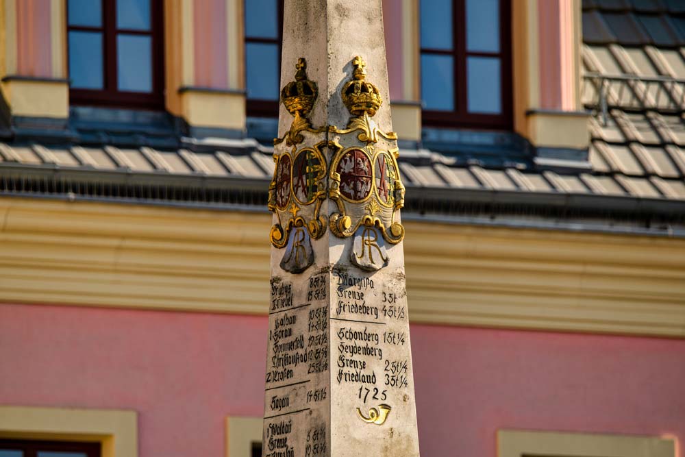 Zbliżenie na górną część Słupa dystansowego poczty polsko-saskiej w Zgorzelcu, z widocznymi herbami i inskrypcjami oraz złotymi koronami na tle różowej kamienicy.