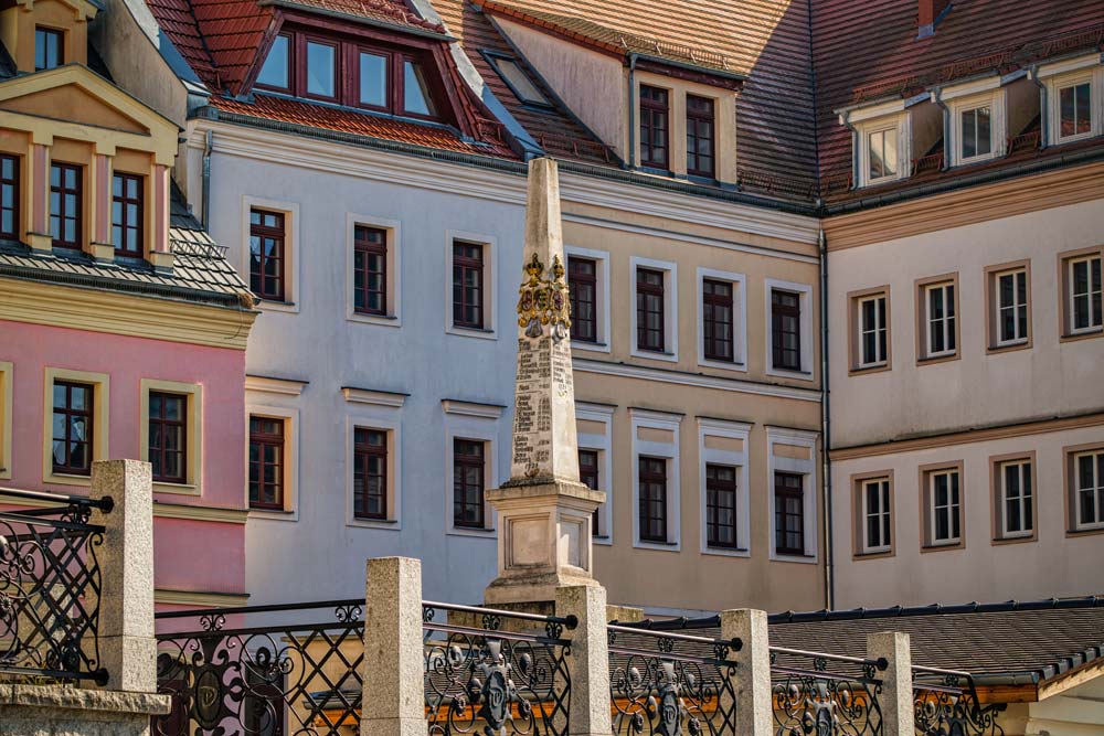 Słup dystansowy poczty polsko-saskiej w Zgorzelcu stojący na placu otoczonym kolorowymi kamienicami, z widoczną metalową balustradą na pierwszym planie.