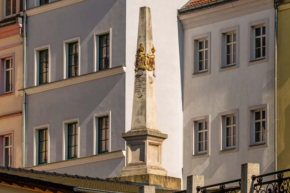 Słup dystansowy poczty polsko-saskiej w Zgorzelcu stojący pomiędzy kamienicami o pastelowych elewacjach, widoczny w pełnej wysokości w popołudniowym słońcu.