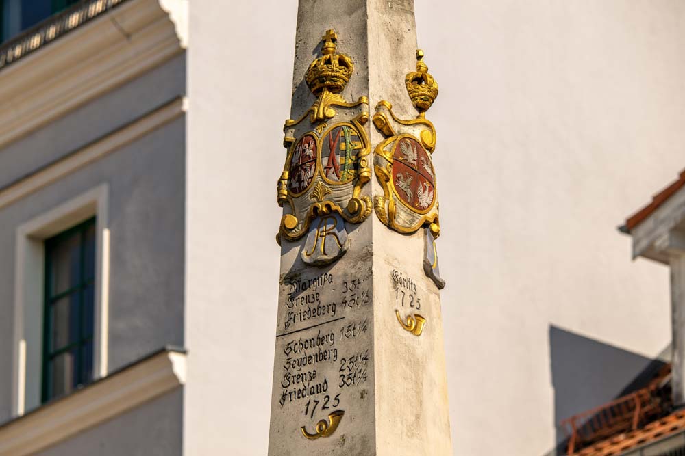 Zbliżenie na inskrypcje i herby na Słupie dystansowym poczty polsko-saskiej w Zgorzelcu, z widoczną datą 1725 i złotymi detalami na tle jasnej elewacji.