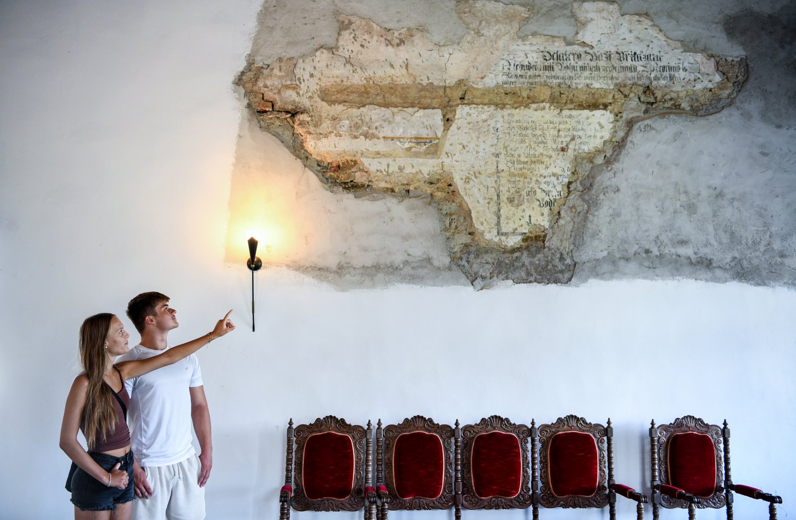 Zwiedzanie wnętrz Kolštejnu – para ogląda odsłonięte fragmenty dawnych malowideł ściennych nad rzędem czerwonych, rzeźbionych krzeseł.