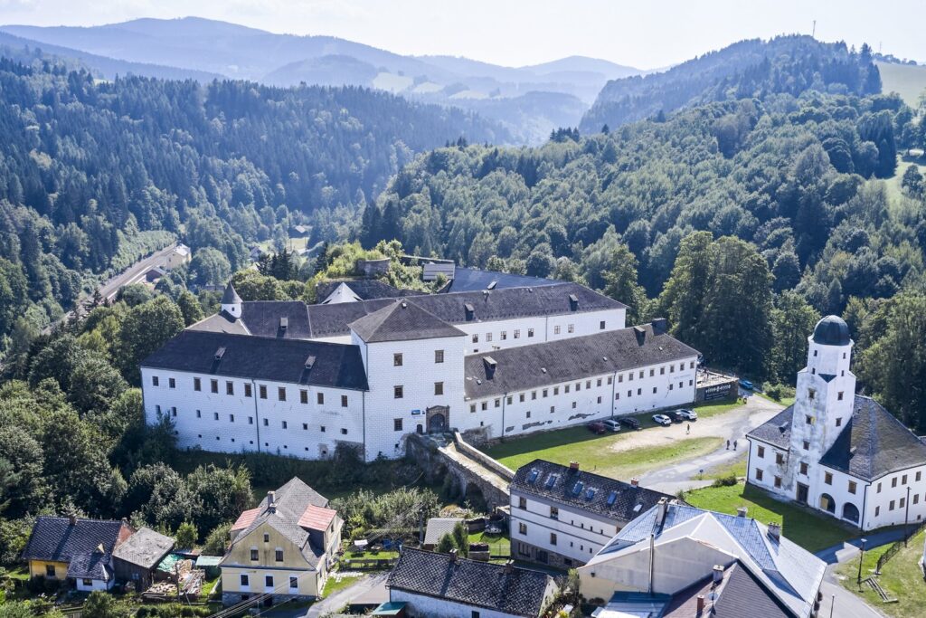 Panorama Pałacu Kolštejn w Brannej – renesansowy zamek na skalnym urwisku pośród zielonych stoków Jesioników, otoczony lasami i wioską.