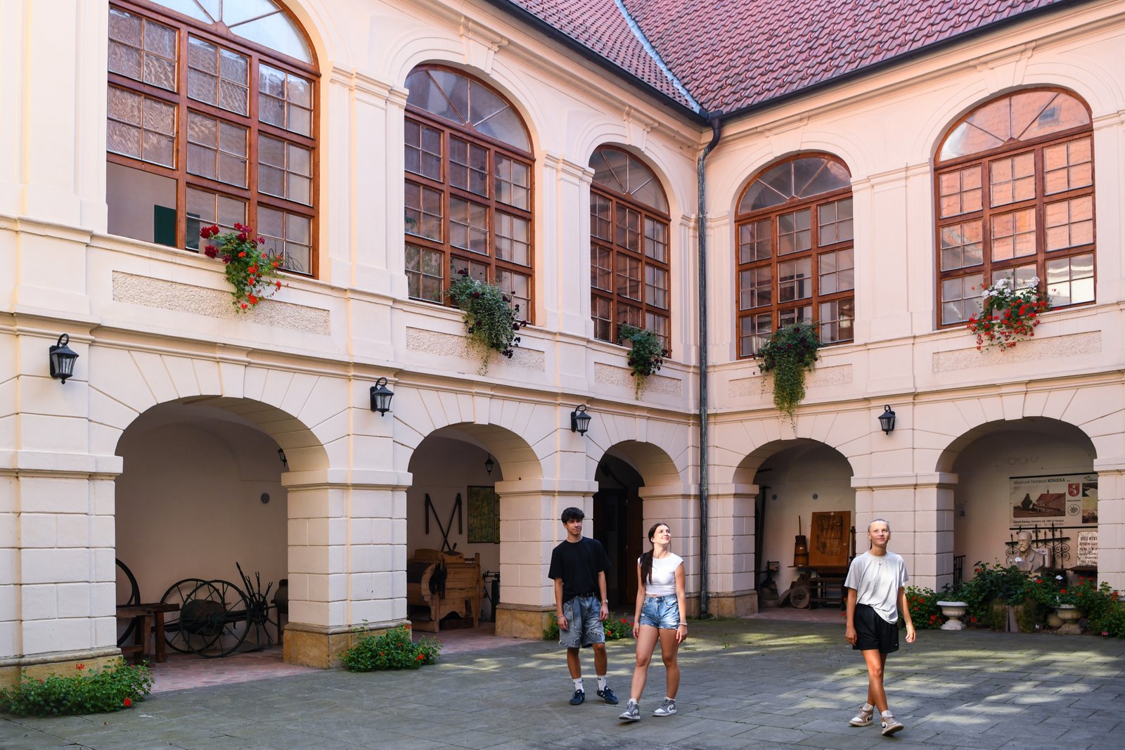 Dziedziniec Pałacu Konice otoczony dwukondygnacyjnymi arkadami z dużymi oknami i kwiatami w skrzynkach, po którym spaceruje trójka młodych turystów.