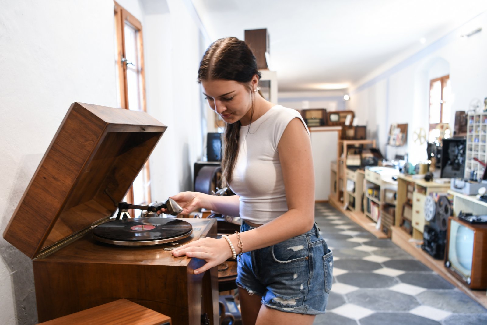 Młoda kobieta obsługuje stary gramofon na korbkę w muzeum rzemiosł w Konicach, otoczona ekspozycją zabytkowych radioodbiorników i sprzętów.