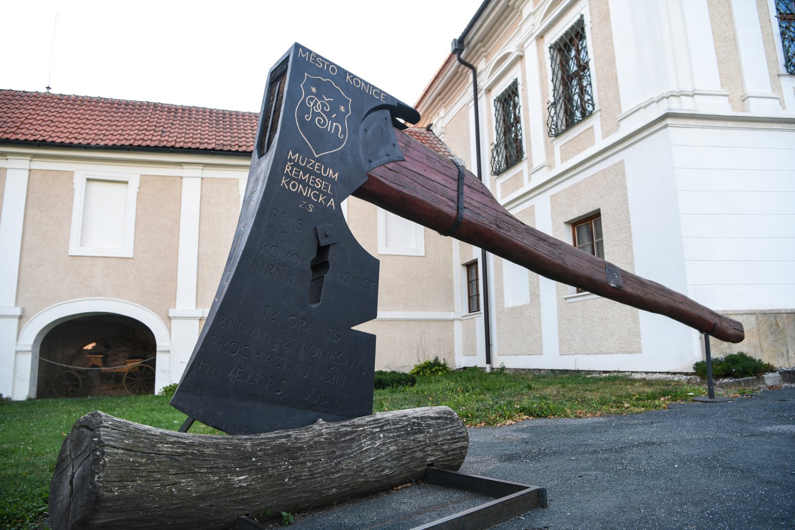 Monumentalna rzeźba topora z długim drewnianym trzonkiem, symbol kolekcji siekier Muzeum Rzemiosł w Konicach, stoi na dziedzińcu barokowego pałacu.