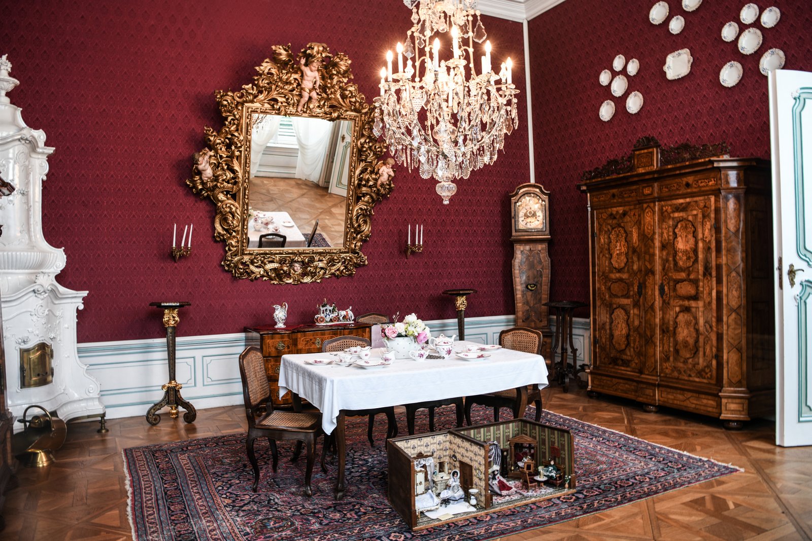 Czerwony salon jadalny w Pałacu Náměšť na Hané – stół przykryty białym obrusem, otoczony krzesłami, na ścianie wielkie złocone lustro, kryształowy żyrandol i masywne drewniane meble na orientalnym dywanie.