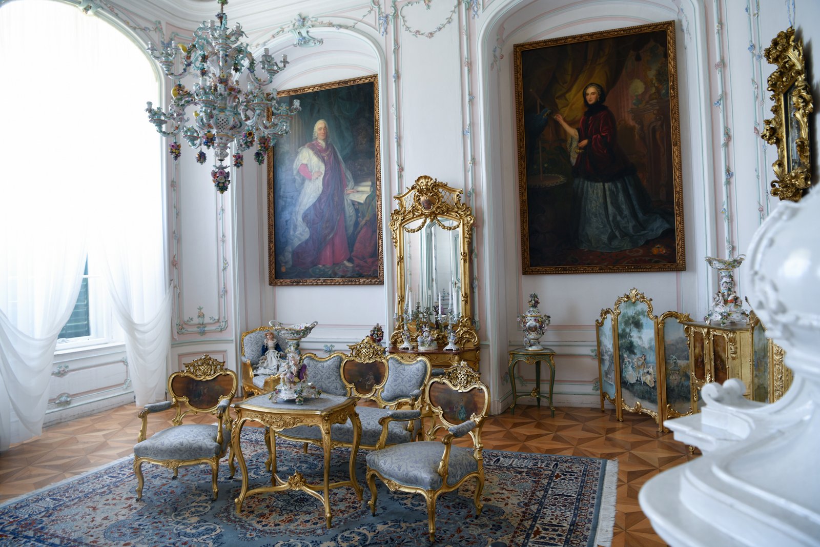 Rokokowy salon w Pałacu Náměšť na Hané – jasne wnętrze z dużymi portretami arystokratów, kryształowym żyrandolem i złoconym zestawem kanapy, foteli oraz stolika na bogato zdobionym dywanie.