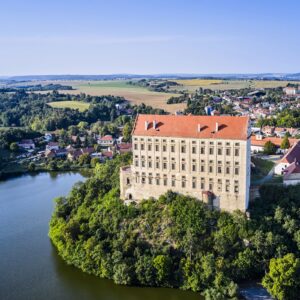 Zdjęcie z drona pokazujące Pałac Plumlov górujący nad jeziorem i miasteczkiem na Morawach; smukła bryła zamku wyraźnie dominuje w krajobrazie.