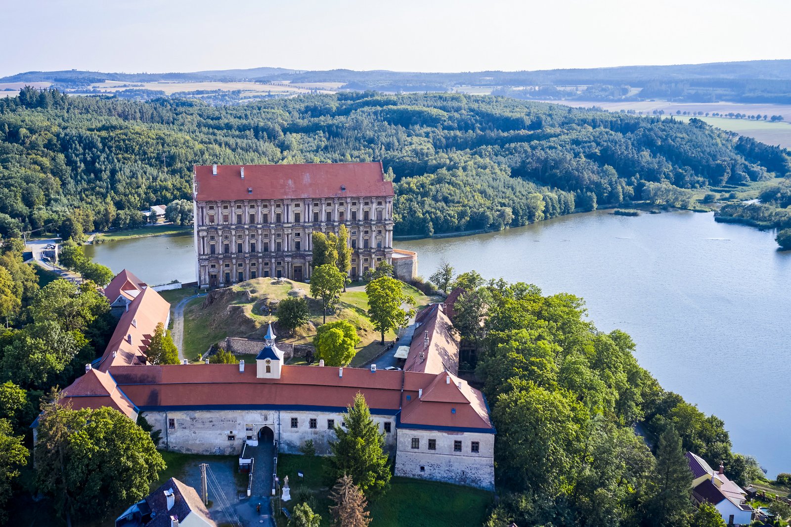 Lotnicze ujęcie kompleksu Pałacu Plumlov z dolnym zamkiem, bastionem i Podhradzkim stawem, otoczonych lasami i wzgórzami regionu Haná. 