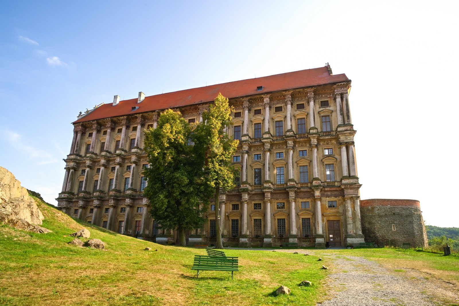 Fasada Pałacu Plumlov na Morawach – wysoka, wąska budowla w stylu wczesnego manieryzmu z czerwonym dachem, stojąca na skarpie nad zielonym trawnikiem.