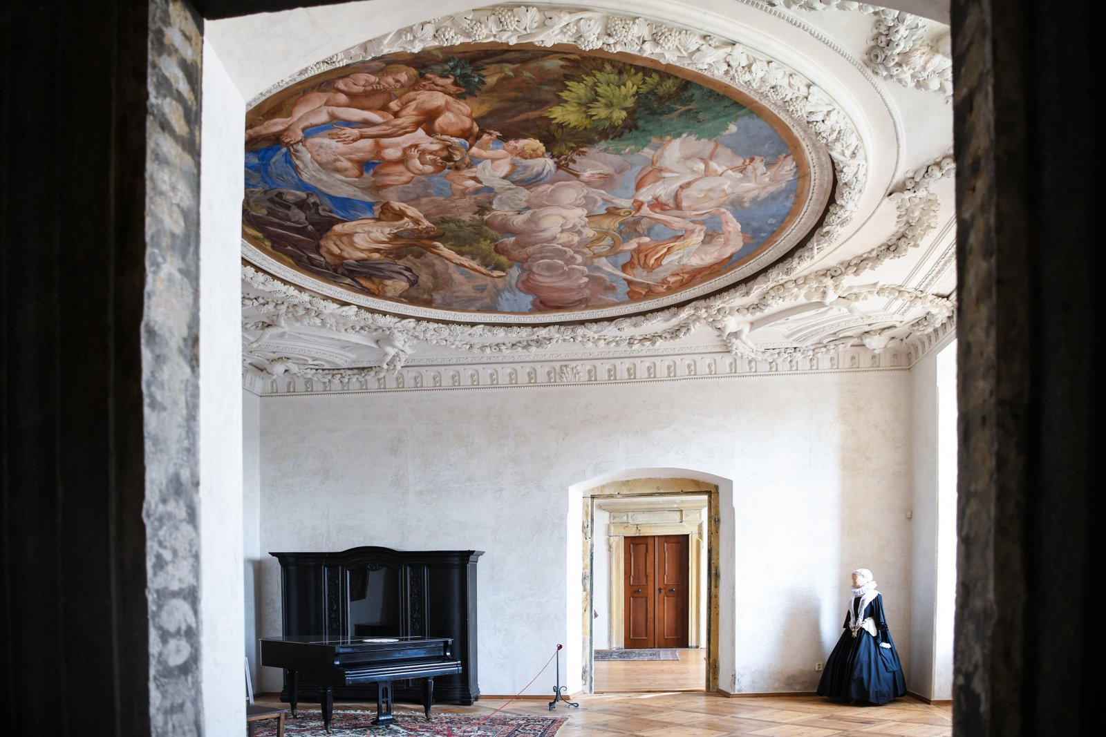 Reprezentacyjna sala Pałacu Plumlov z barokowym malowidłem na owalnym sklepieniu, bogatą sztukaterią, czarnym fortepianem i manekinem w historycznym stroju.