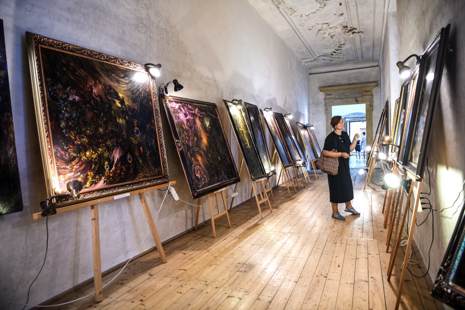 Wnętrze Pałacu Plumlov – długi korytarz zamieniony w galerię sztuki z dużymi obrazami na sztalugach, oświetlonymi lampami, i kobietą oglądającą wystawę.