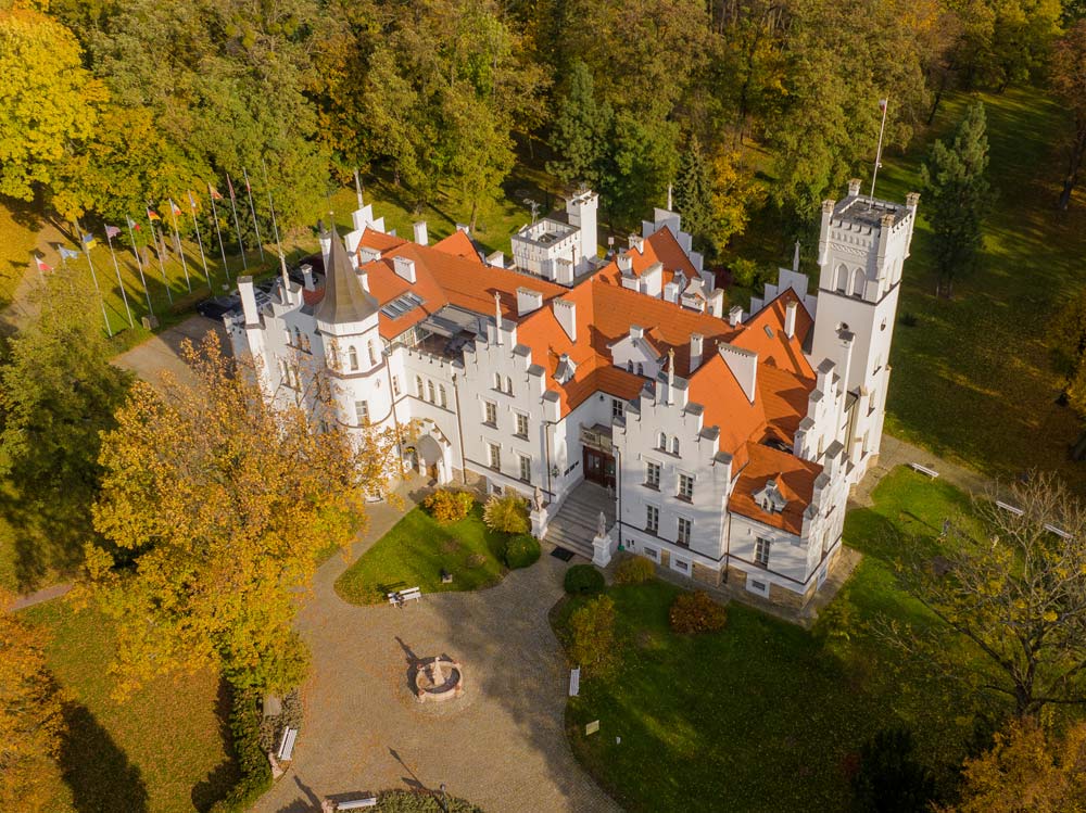 Ujęcie z drona na kompleks Pałacu Sulisław z czerwonym dachem pośrodku zielonego parku, z widoczną fontanną i alejkami przed frontem