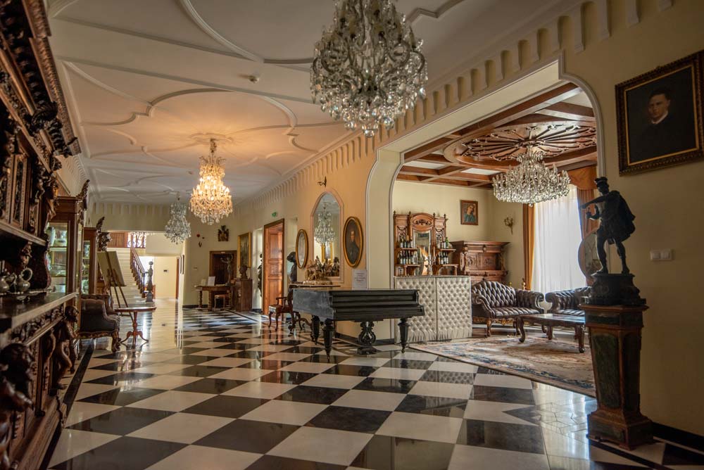 Reprezentacyjny hol Pałacu Sulisław z połyskującą szachownicową posadzką, kryształowymi żyrandolami, zabytkowymi meblami i salonem z sofami po prawej stronie.