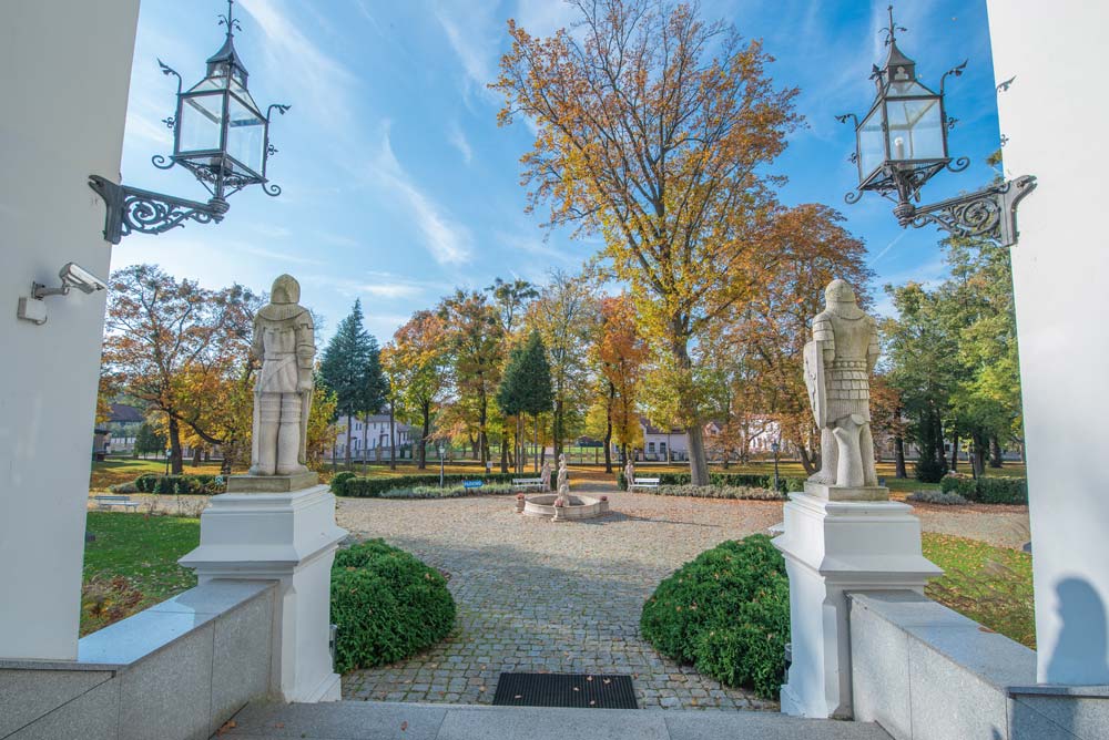 Widok z pałacowych schodów na jesienny park przed Sulisławiem, z dwiema kamiennymi figurami rycerzy po bokach wejścia i fontanną pośrodku alejek.