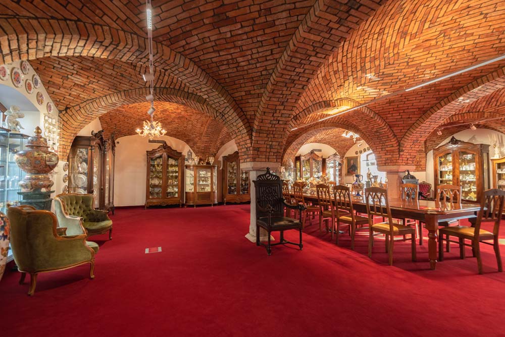 Rozległe, ceglane wnętrze pałacowych piwnic z długim stołem i krzesłami, zielonymi fotelami oraz rzędami przeszklonych witryn z porcelaną na czerwonej wykładzinie.
