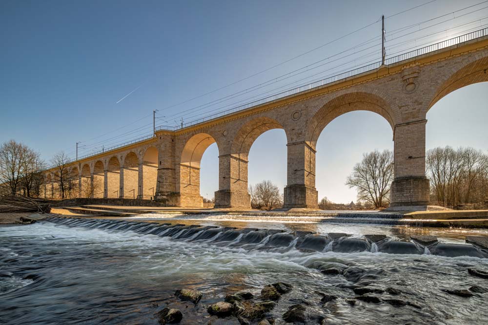 Wiadukt kolejowy w Bolesławcu widziany znad bystrza na Bobrze – kamienne arkady i płynąca woda na pierwszym planie.