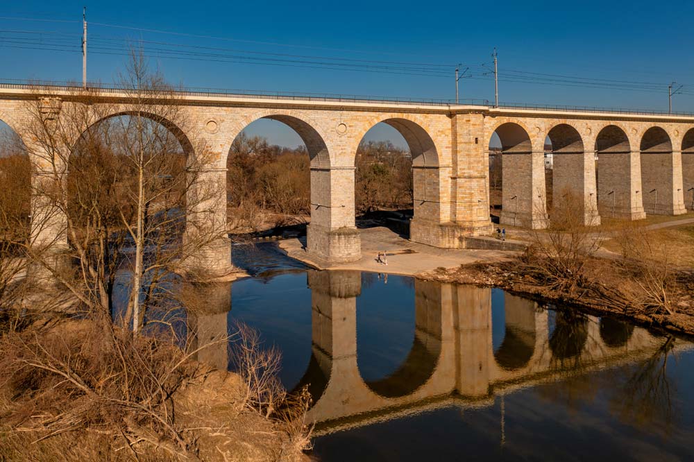 Fragment wiaduktu kolejowego w Bolesławcu – wysokie łuki odbijające się w spokojnej tafli rzeki.