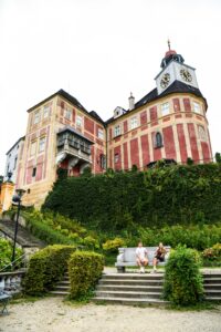 Zewnętrzne ujęcie pałacu Jánský Vrch z dołu: masywne, różowo-żółte mury z wieżą zegarową wyrastają ponad zieloną skarpę porośniętą roślinnością, a na schodach w dolnej części kadru siedzi para turystów odpoczywająca na kamiennej ławce.