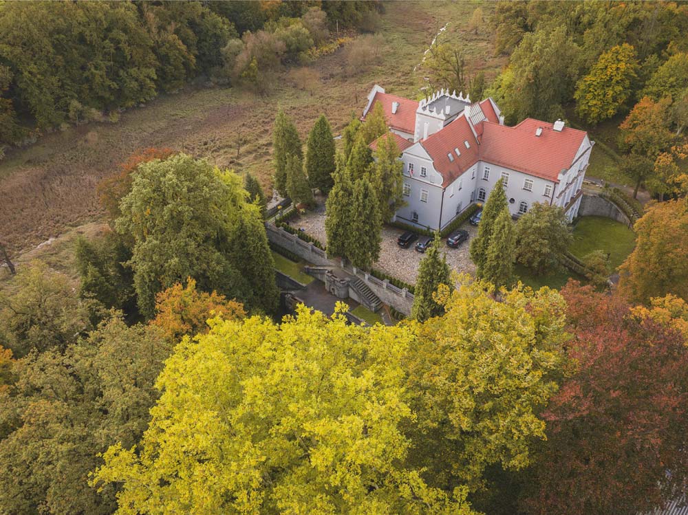 Zbliżenie z lotu ptaka na zabudowania zamku w Rogowie Opolskim – biały budynek z czerwonym dachem otoczony koronami drzew.