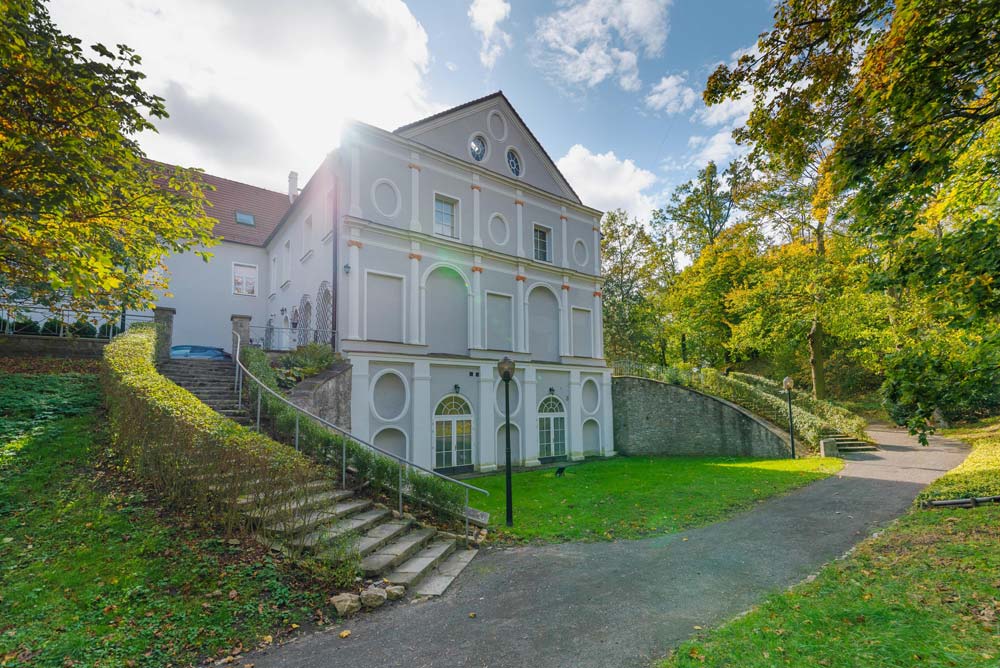 Elewacja zamku w Rogowie Opolskim od strony parku: jasny budynek, schody, mur oporowy i alejka biegnąca wśród zieleni.