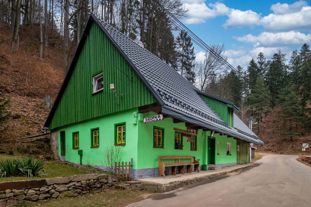 Widok z ukosa na zieloną chałupę ruchomej szopki przy wąskiej drodze; ciemny, stromy dach, las w tle i znak „Szopka ruchoma” przy ganku.