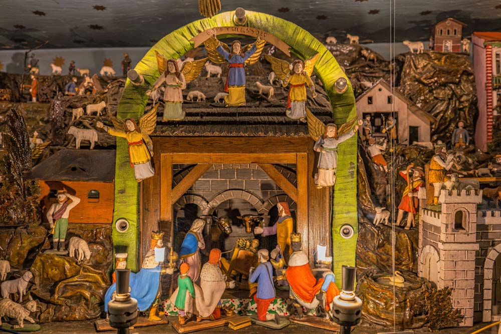 Zbliżenie na scenę Bożego Narodzenia w ruchomej szopce: stajenka pod zielonym łukiem z aniołami, a przed nią postacie Trzech Króli i pasterzy.
