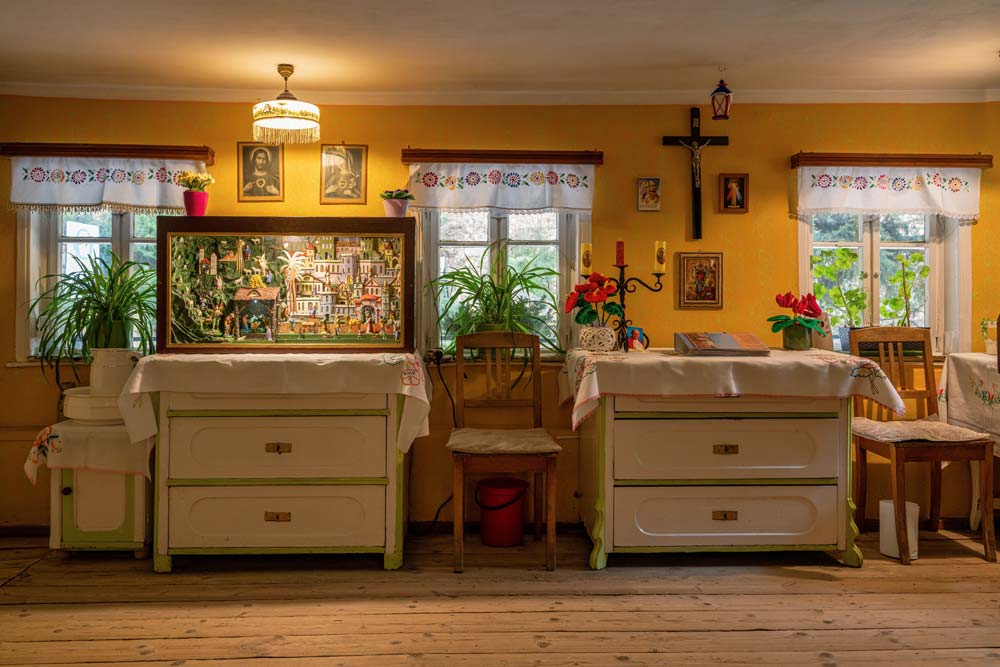 Przytulne wnętrze chałupy w Czermnej: stoły z białymi obrusami, rośliny w oknach, krucyfiks na ścianie i ekspozycja ruchomej szopki na jednym z blatów.