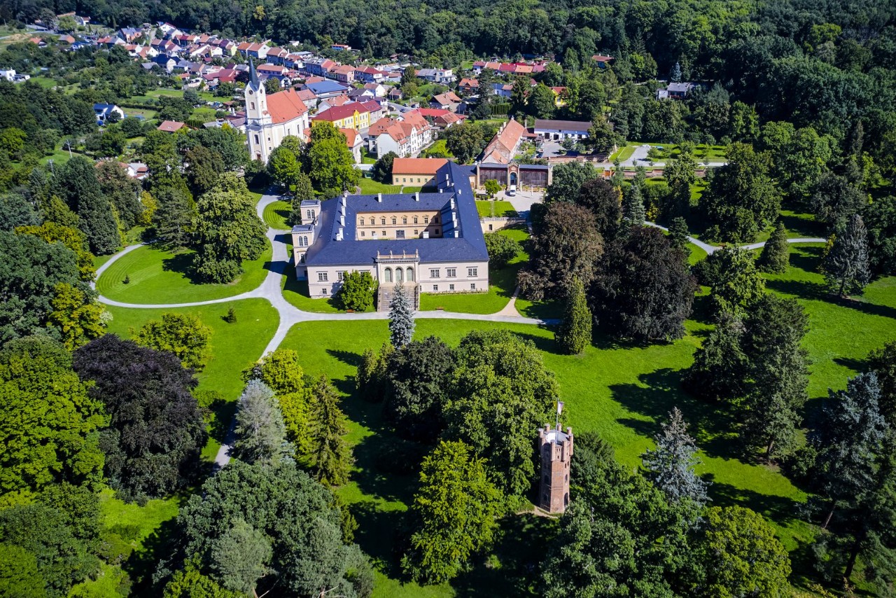 Widok z lotu ptaka na zespół pałacowo-parkowy Čechy pod Kosířem: klasycystyczny pałac otoczony rozległym parkiem pełnym drzew, a wśród zieleni wyrasta ceglana Czerwona Wieża; w tle miasteczko i ściana lasu.