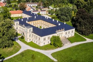 Zbliżony widok z góry na klasycystyczny pałac w Čechach pod Kosířem – czteroskrzydłową rezydencję z wewnętrznym dziedzińcem, ciemnym dachem i monumentalnymi schodami prowadzącymi do otaczającego ją parku.