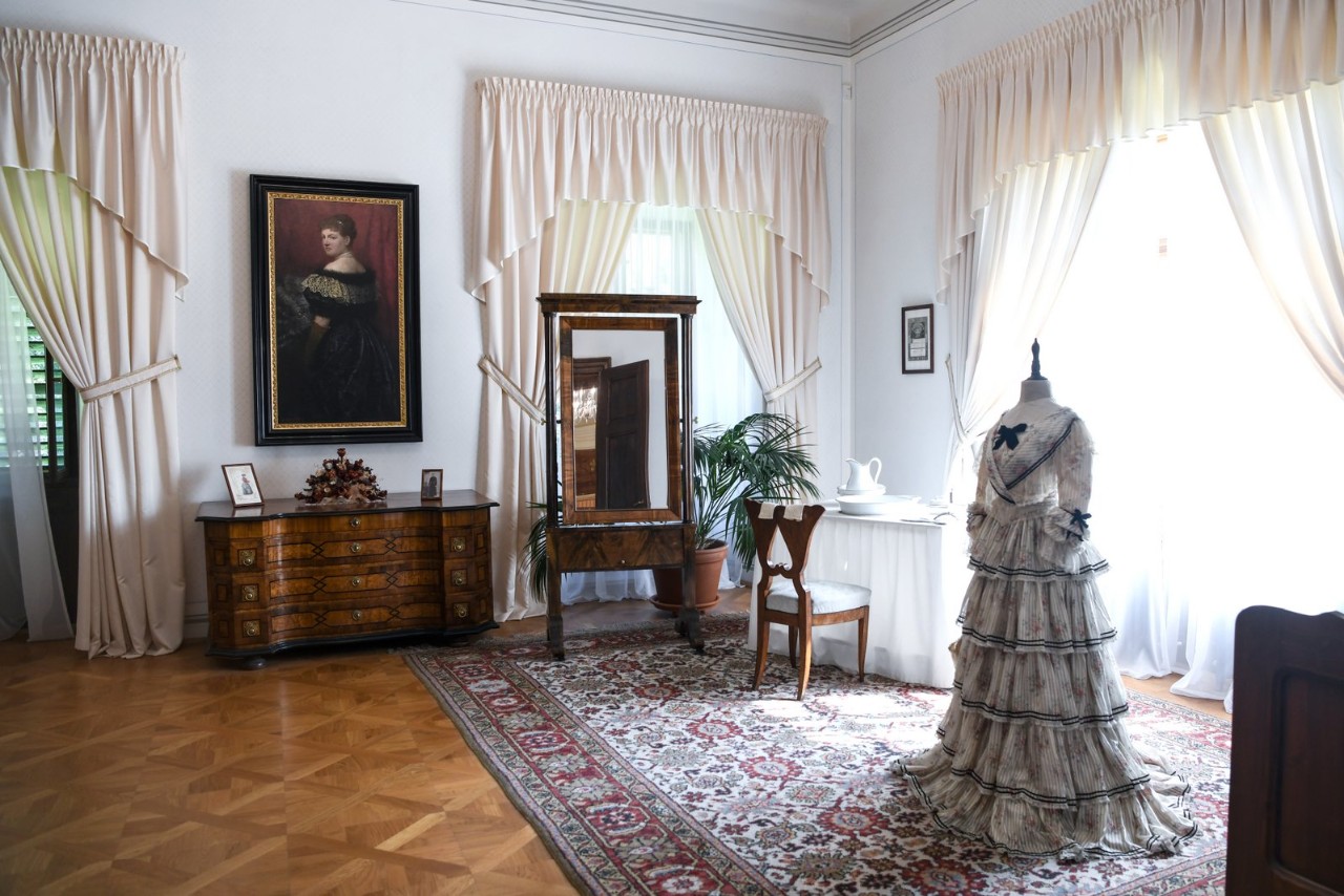 Jasny pokój w pałacu z białymi zasłonami, parkietem i dywanem; po lewej stronie komoda i portret arystokraty, po prawej manekin w długiej, kremowej sukni z falbanami, obok biurko i toaletka.