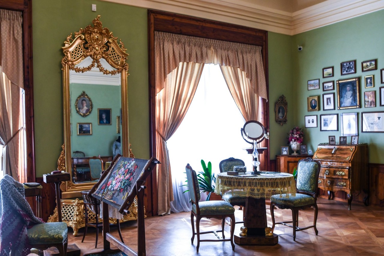 Zielony salon z wysokimi oknami i ciężkimi zasłonami: pośrodku stoi okrągły stół z haftowanym obrusem, wokół krzesła, z lewej strony złocone lustro w bogatej ramie, a na ścianie wisi galeria rodzinnych fotografii.