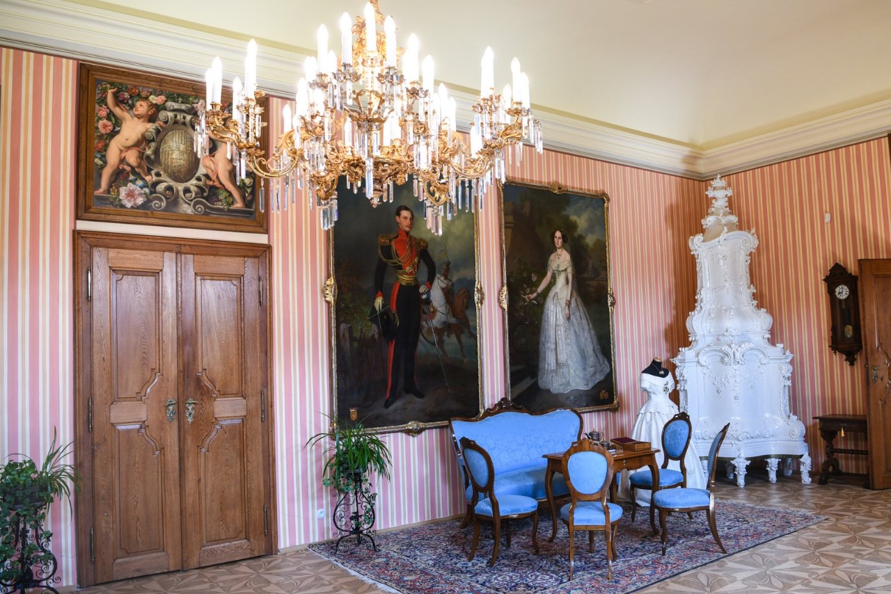 Reprezentacyjny salon pałacu Čechy pod Kosířem z różowo-pasiastymi ścianami, wielkim kryształowym żyrandolem, błękitnym kompletem wypoczynkowym, białym piecem kaflowym i portretami arystokratów w złotych ramach.