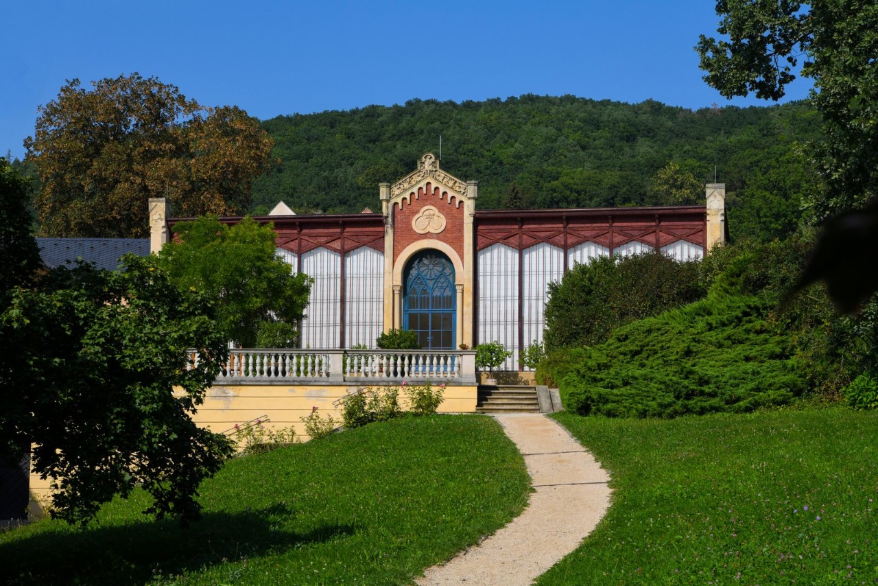Ścieżka prowadzi przez zielony trawnik do neogotyckiej oranżerii Pałacu Čechy pod Kosířem – przeszklonej fasady z ozdobnym szczytem, na tle zalesionego wzgórza Kosíř i błękitnego nieba.