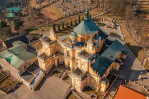 Widok z drona na sanktuarium w Wambierzycach: żółto-biała bazylika z zielonym dachem i rozległymi schodami, otoczona zabudowaniami.