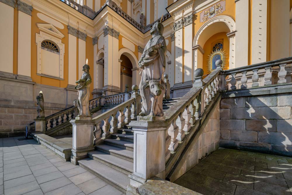 Kamienne schody i balustrady przy bazylice w Wambierzycach, ozdobione figurami świętych na postumentach, w ostrym świetle popołudnia.