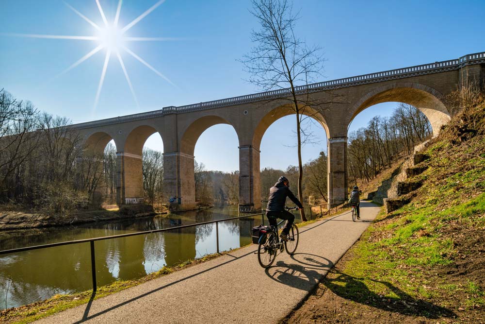 Ścieżka rowerowa wzdłuż Nysy Łużyckiej pod kamiennymi łukami mostu; dwóch rowerzystów jedzie w słońcu przy brzegu rzeki.
