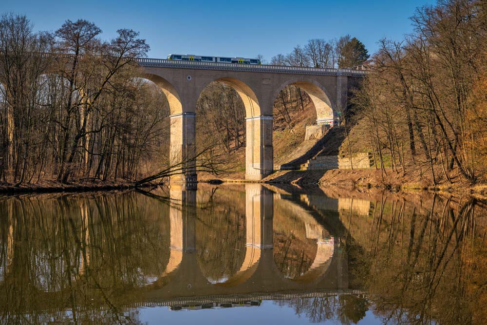 Kamienny most kolejowy nad Nysą Łużycką odbity w spokojnej wodzie; na górze przejeżdża pociąg między Zgorzelcem a Görlitz.