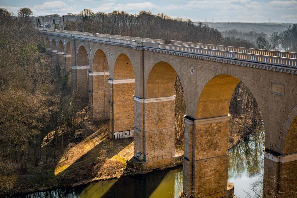 Zbliżenie na zabytkowy, kamienny most kolejowy nad Nysą Łużycką w Zgorzelcu – długie łuki nad rzeką i zalesioną doliną.