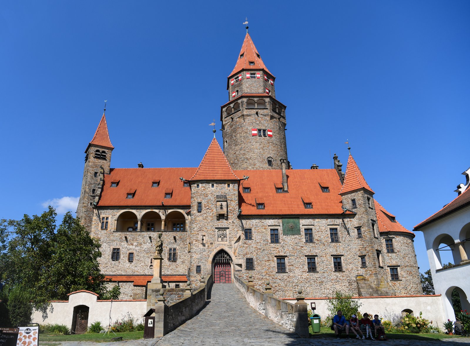 Główna fasada i wjazd – kamienny most prowadzący do bramy pod wysoką wieżą, w słoneczny dzień z czystym niebem.