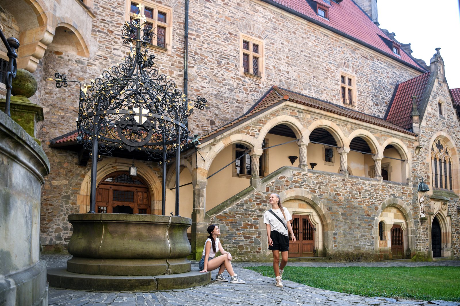 Dziedziniec z ozdobną studnią – ażurowa, żelazna konstrukcja nad cembrowiną i arkadowe krużganki w tle; dwie osoby odpoczywają przy studni.