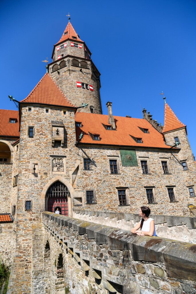 Widok na wieżę i bramę od strony mostu – kamienne mury i czerwone dachy, z postacią opartą o balustradę w pierwszym planie.