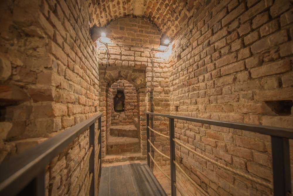 Ceglane podziemia – wąski korytarz ze sklepieniem z cegły, oświetlony ciepłym światłem; po bokach metalowe barierki z liną, na końcu małe przejście i wnęka w murze.