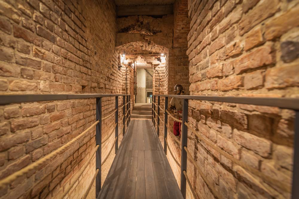 Podziemny pomost – dłuższe ujęcie tego samego typu przejścia: drewniany/metalowy pomost prowadzi przez ceglaną przestrzeń do otwartego przejścia; po prawej stoi manekin/figura w stroju