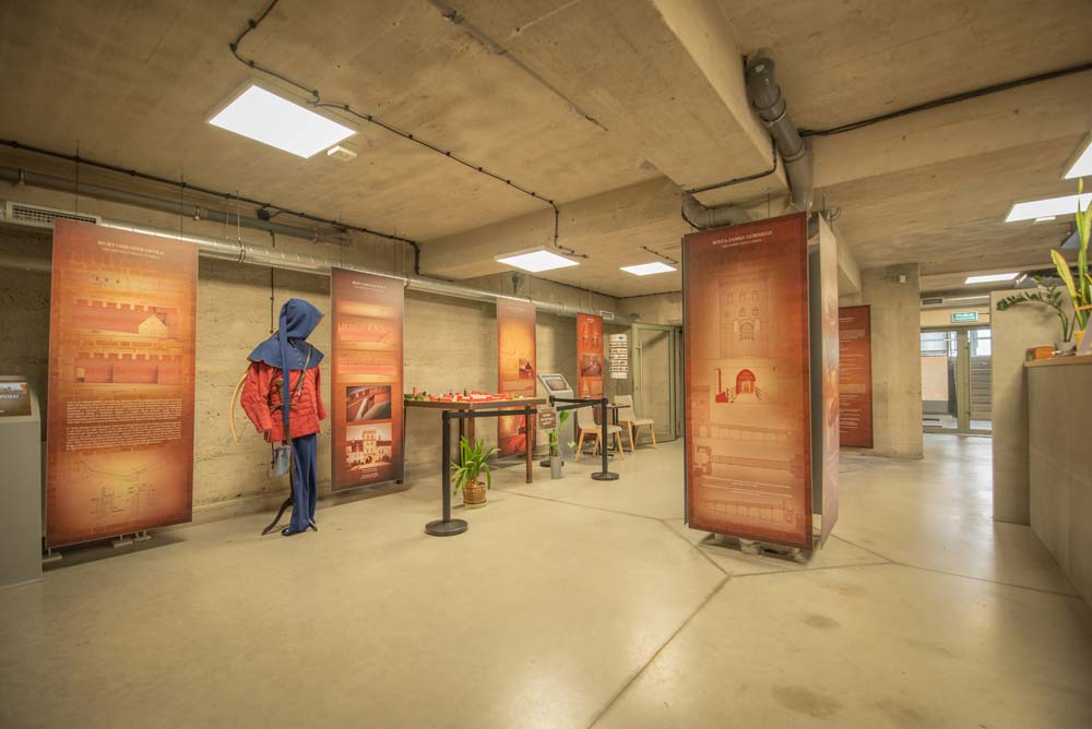 Nowoczesna sala ekspozycyjna – surowy betonowy sufit i słupy, duże stojące plansze z opisami, makieta na stoliku; po lewej manekin w czerwonym stroju z niebieskim „kapturem”/nakryciem głowy