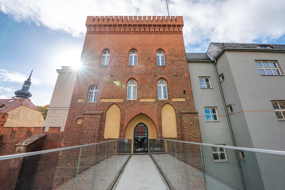 Wejście po szklanym moście – perspektywa „na wprost” na ceglaną fasadę z ostrołukowym portalem; słońce daje flarę w lewym górnym rogu, po bokach szkło balustrad.