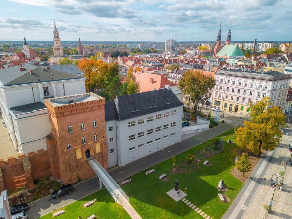 Panorama miasta z lotu ptaka – kompleks wieży na pierwszym planie, dalej gęsta zabudowa Opola i charakterystyczne kościoły (widoczne wieże), jesienne drzewa i szeroki horyzont.