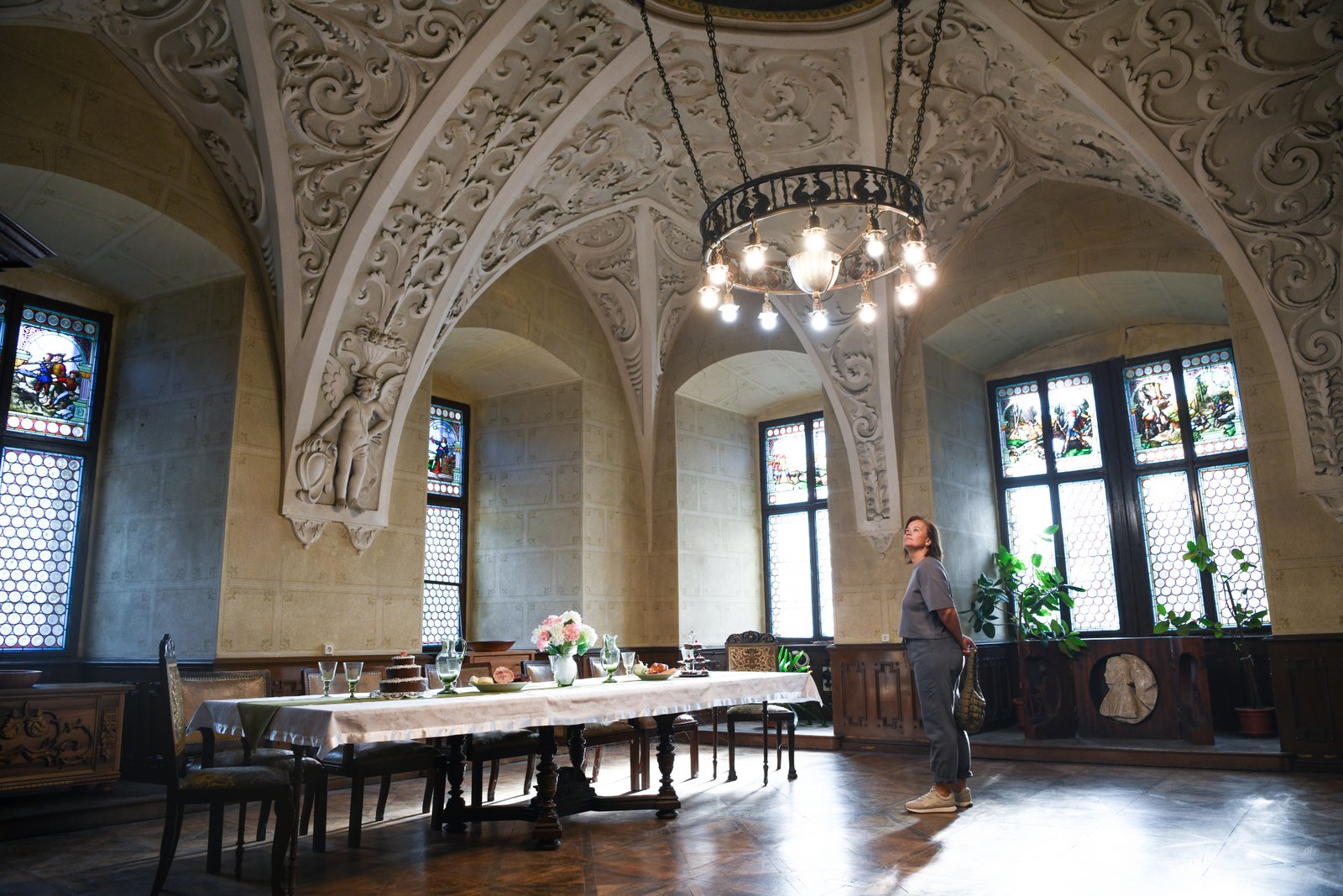 Reprezentacyjna sala z dekoracyjnym sklepieniem – bogato rzeźbione żebra i ornamenty, witraże w oknach, długi stół nakryty obrusem; zwiedzająca patrzy w górę.